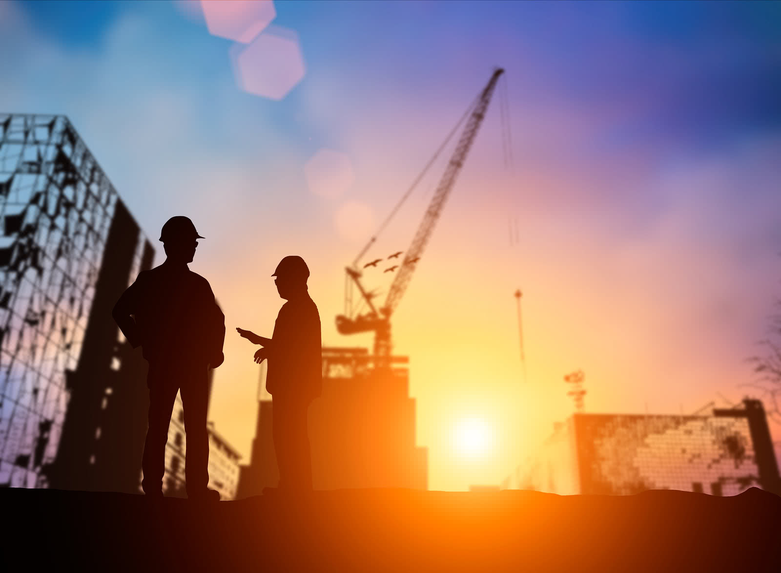 Two construction workers stand in front of a crane with sunset background