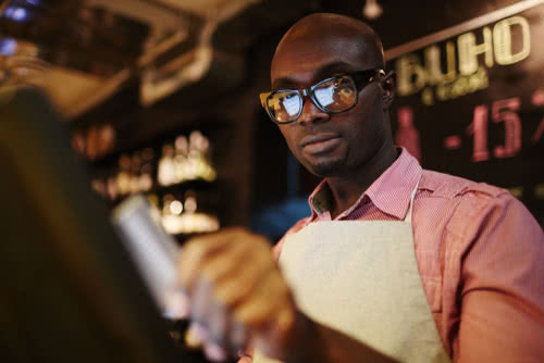 A male waiter in glasses uses a POS system while working in restaurant