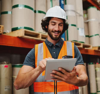 Male warehouse manager adding stock inventory data in digital tablet in warehouse wearing a white hardhat and safety vest standing