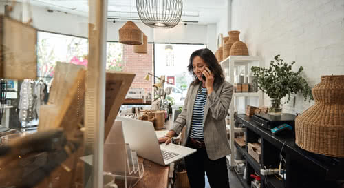 Smiling young woman standing behind a counter in her stylish boutique working on a laptop and talking on a cellphone